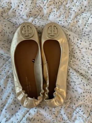 Guldiga ballerina från Tommy Hilfiger - Säljer ett par flexibla ballerina i metallic guld från Tommy Hilfiger. Skorna har rund tå, elastisk kant och en stor guldfärgad logga framtill. 