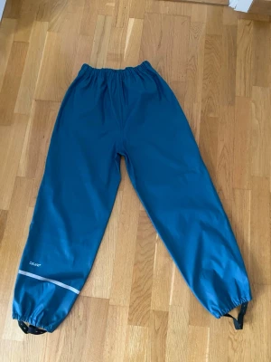Blå täckbyxor Everest  - Blå täckbyxor från everest med resår i midjan och benslut. Byxorna har en reflexdetalj på ena benet och justerbara band vid anklarna. Perfekta för utomhusaktiviteter under kyliga dagar.