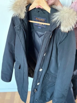 Svart jacka Woolrich med pälskant - Säljer en svart jacka från Woolrich med stor huva och fluffig pälskant. Jackan har dragkedja och knappar framtill, två stora fickor och en klassisk passform. Perfekt för kalla dagar och riktigt snygg till alla outfits. Storlek XS.