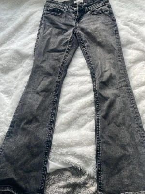 Grå bootcut jeans med slitna detaljer - Säljer ett par grå jeans med bootcut-modell och snyggt utsvängda ben. Jeansen har klassisk femficksdesign och är tillverkade i jeansmaterial. Färgen är en tvättad grå nyans och nederkanten har slitna detaljer för en cool look. Ändan av benen är lite slitna