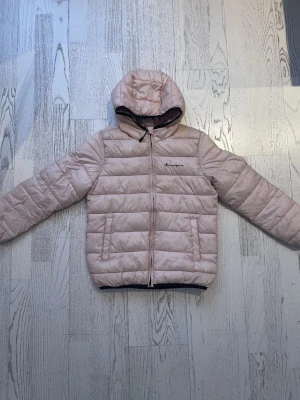 Ljusrosa pufferjacka från Champion - Säljer en ljusrosa pufferjacka från Champion med huva och dragkedja framtill. Jackan har quiltad design, två fickor och Champion-logga broderad på bröstet. Insidan är mönstrad med Champion-tryck. Perfekt för kyliga dagar. Lite smutsig på ärmarna bara!