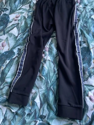 Svarta mjukisbyxor från Hugo Boss M - Svarta mjukisbyxor från Hugo Boss med vita ränder längs sidorna och diskret Boss-logga på benet. Byxorna har resår i midjan och mudd vid bensluten. Tillverkade i mjukt syntetmaterial och har en sportig look med snörning i midjan.