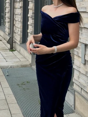 Marinblå sammetsklänning offshoulder - Säljer en marinblå långklänning i mjuk sammet med offshoulder-design och v-ringad omlott upptill. Klänningen har en elegant slits framtill och smickrande drapering i midjan. Perfekt för dig som vill ha en stilren och lyxig look.