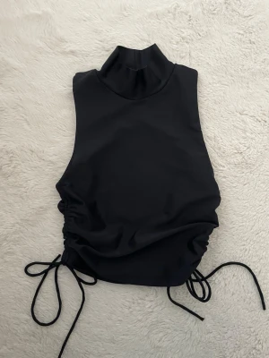 Svart polotopp med snörning i sidan - Säljer en svart ärmlös polotopp med hög krage och snygg snörning i sidorna som ger en cool rynkad effekt. Toppen är croppad och har en stilren, minimalistisk look. Perfekt att styla med jeans eller kjol för en trendig outfit. 