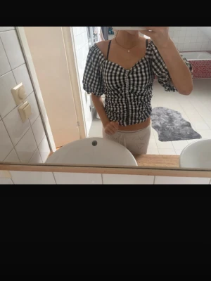 Svartvit rutig blus med puffärm - Säljer en svartvit rutig blus med puffiga korta ärmar och smockad midja. Toppen har en söt v-ringning och sitter croppad på kroppen. Perfekt för dig som gillar trendiga detaljer och vill ha en topp som sticker ut.