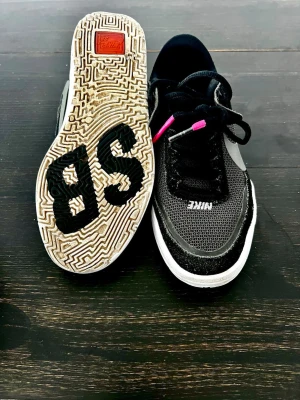 Nike SB Day One sneakers svart och grå. - Nike SB sneakers i svart och grå mesh med vita detaljer och rosa accenter på snörningen. Skorna har en vit sula med SB-logga undertill och klassisk Nike swoosh på sidan. Ovandelen är i mesh och mocka för en sportig look.