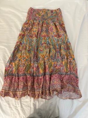 Färgglad långkjol från Marta - Säljer en bohemisk långkjol från Marta med färgstarkt blommigt och paisleyinspirerat mönster i rosa, gul, blå och gröna toner. Kjolen har bred smock i midjan och volangdetaljer längst ner. Perfekt för dig som gillar unika prints och sköna material.