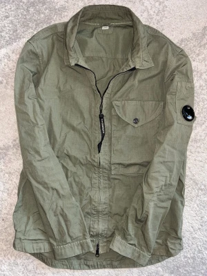Grön overshirt från C.P. Company - Snygg grön overshirt från C.P. Company med dragkedja och knappstängd bröstficka. Ikonisk linsdetalj på ärmen. Väldigt tunt o luftigt material, perfekt lager-på-lager-plagg med modern känsla. Strl M men har du S så funkar de på dig också. Priset är prutbart. Hör av er vid intresse/ frågor!