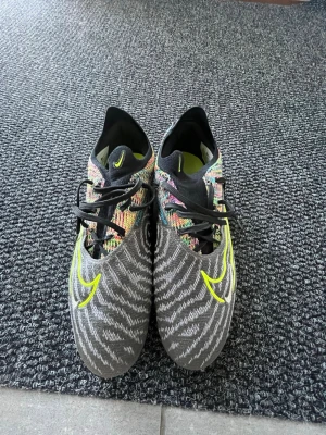 Nike phantom gx  - Säljer ett par Nike fotbollsskor med grå bas och stickat mönster i vitt, samt färgglada detaljer i gult, rosa och blått. Skorna har snörning, rund tå och är tillverkade i syntetmaterial med stickad ovandel för extra komfort och passform. Har använt dessa inte fler än 5 gånger, absolut inget fel på dem.