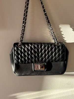 Svart quiltad axelväska Karl Lagerfeld - Snygg svart axelväska från Karl Lagerfeld med quiltad ovandel och kedjerem i silver. Väskan har ett klassiskt rektangulärt format, vridlås framtill och detaljer i metall. Insidan är fodrad med mönstrat tyg och har flera fack för småsaker.