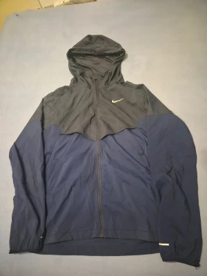 Nike miller jacka - Säljer en snygg tvåfärgad vindjacka från Nike i mörkblått och grått. Jackan har huva med dragsko, hel dragkedja framtill och reflekterande detaljer. Perfekt för blåsiga dagar och har meshfoder på insidan för extra komfort.