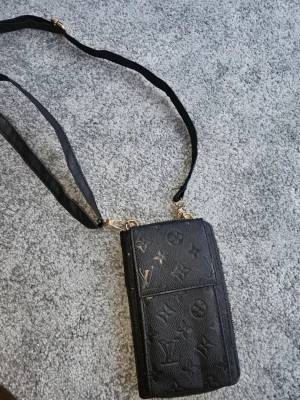 Svartaxelväska i skinn - Säljer en svart axelväska i präglat skinn med klassiskt monogrammönster. Väskan har justerbar axelrem, guldfärgade detaljer och flera fack med dragkedja för kort och småsaker. Perfekt storlek för mobilen och det viktigaste.