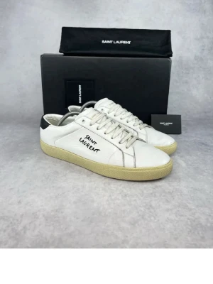 Saint Laurent vita sneakers med svart detalj - Säljer ett par snygga vita sneakers från Saint Laurent med handskriven logga på sidan. Skorna har svart detalj vid hälen, beige gummisula och är tillverkade i skinn. Klassisk rund tå och snörning framtill. Perfekta för dig som gillar stilrena och trendiga sneakers. Kan sälja dem för billigare vid en snabb och smidig affär.