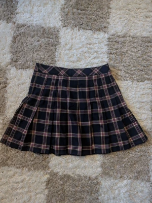 Svart rutig plisserad kortkjol - Säljer en svart plisserad kortkjol med rutigt mönster i vinrött, beige och vitt. Kjolen har en klassisk midja och stängs med dragkedja och knapp i sidan. Perfekt för dig som gillar preppy stil och vill ha något som sticker ut.