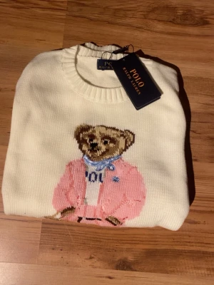 Vit stickad tröja Polo Ralph Lauren - Vit stickad tröja från Polo Ralph Lauren med klassisk björn broderad på framsidan i rosa outfit och blå scarf. Tröjan har rund hals, ribbade muddar och RL-broderi vid nederkanten. Perfekt för dig som gillar ikoniska detaljer och snygg design.