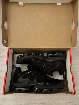 Nike Air Vapormax Plus Svart 43 - Nike Air Vapormax Plus sneakers i helsvart med mörkgrå detaljer. Snygg design med synlig Air-sula och vågiga TPU-paneler på ovansidan. Skorna har snörning och platt sula med rund tå. Materialet är syntet och skinn, perfekt för dig som gillar sportig stil. Levereras i originalboxen.