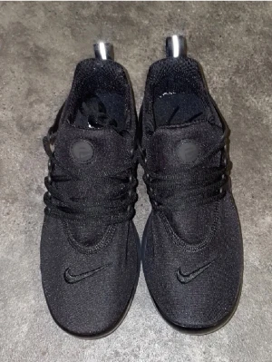 Svarta Nike Presto sneakers - Säljer ett par helsvarta Nike Presto sneakers med klassisk snörning och rund tå. Skorna har en stilren design med diskret Nike-logga på tån och är tillverkade i ett mjukt syntetmaterial som ger en skön passform. Perfekta för dig som gillar en clean och sportig look.