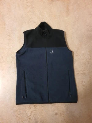 Svart och mörkblå fleeceväst Haglöfs - Mysig fleeceväst från Haglöfs i svart och mörkblått. Västen har hög krage, två fickor med dragkedja och diskret Haglöfs-logga på bröstet. Perfekt lager för kyliga dagar och outdoor-häng.