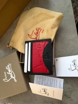 Christian Louboutin svart och röd plånbok - Säljer en snygg plånbok från Christian Louboutin i svart skinn med röd detalj och präglad logga på framsidan. Plånboken har flera kortfack och stilren design, perfekt för dig som vill ha något både lyxigt och praktiskt. Helt nytt.