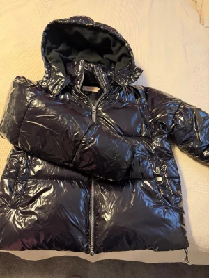 Svart glansig pufferjacka från Nelly - Säljer en svart glansig pufferjacka från Nelly i storlek S. Jackan har huva, dragkedja framtill och två fickor med knappar. Perfekt för kalla dagar och riktigt snygg med sin blanka finish. Passar dig som gillar en lite oversized vibe.