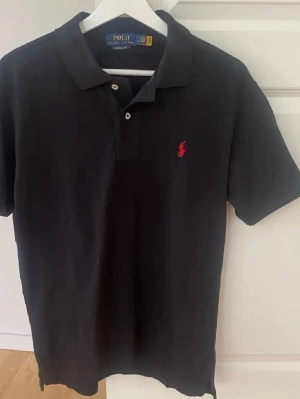 Svart pikétröja från Polo Ralph Lauren - Klassisk svart pikétröja från Polo Ralph Lauren med röd broderad logga på bröstet. Tröjan har korta ärmar, krage och två knappar framtill. Tillverkad i bomull för en skön och luftig känsla. Perfekt till jeans eller shorts.