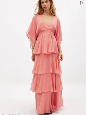 Rosa långklänning med volanger - Supersöt långklänning i ljusrosa med flera lager volanger och v-ringning. Klänningen har vida, korta ärmar och omlottliv som ger en flowy känsla. Perfekt för sommarens alla tillfällen och ger en romantisk vibe. Materialet känns lätt och luftigt. Använt en gång.