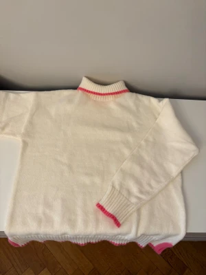 Vit stickad tröja med rosa detaljer - Vit stickad tröja från MEET ME THERE i storlek XXS/XS. Tröjan har ribbade muddar och polokrage med rosa kant, samt rosa vågiga detaljer vid ärmslut och nederkant. Perfekt för dig som gillar söta och stilrena plagg. Aldrig använd