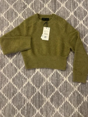 Olivgrön croppad stickad tröja XS - Snygg olivgrön croppad stickad tröja i storlek XS. Tröjan har bred ribbad mudd i midjan och vid ärmsluten, samt rund halsringning. Perfekt för lager-på-lager och chill höst/vinterstil.