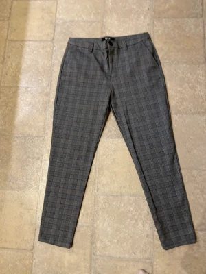 Grå rutiga kostymbyxor från Smog - Snygga grå kostymbyxor från Smog i storlek M. Byxorna har ett rutigt mönster i diskreta toner och klassisk rak passform med fickor i sidorna. Perfekta för en stilren look och funkar till många olika outfits.