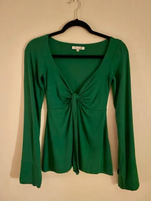 Grön omlottopp från Design by Si - Snygg grön omlottopp från Design by Si i storlek XS/S. Toppen har lång ärm, v-ringning och en knytning framtill som ger en söt detalj tillsammans med passande trumpetärmar. I nytt skick, enbart testad.