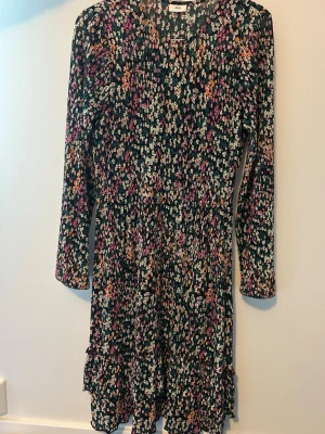 Blommig kortklänning från JDY - Supersöt kortklänning från JDY med blommigt mönster i rosa, orange, vit och grön på mörk botten. Klänningen har lång ärm, markerad midja och volangdetaljer längst ner. Perfekt för dig som gillar färg och mönster!