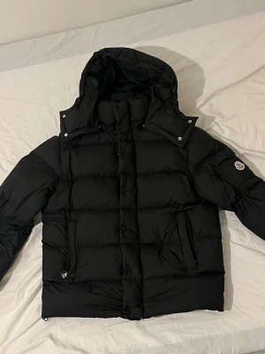 Svart Moncler pufferjacka med huva - Säljer en svart pufferjacka från Moncler med avtagbar huva och klassisk logga på ärmen. Jackan har dragkedja och knappar framtill, två sidofickor och quiltad design. Insidan har Monclers ikoniska serietidningsmärke. Perfekt för kalla dagar.