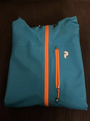 Blå vindjacka Peak Performance XL - Säljer en blå vindjacka från Peak Performance i storlek XL. Jackan har huva med snörning, orange dragkedja och bröstficka med logga. Tillverkad i lätt och slitstark polyester, perfekt för utomhusaktiviteter. Snygg och funktionell design.
