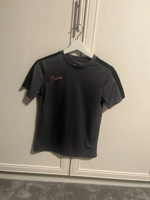 Grå Nike träningströja  - Bilderna visar en svart Nike Dri-Fit t-shirt med rosa logga, perfekt till träning eller vardag skön och bekväm tröja med luftat material. Skulle säga att den är större i storleken så sitter som XS mått finns bara att höra av sig! 