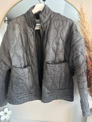 Grå mönstrig jacka från Zara - Grå jacka från Zara med vågigt mönster, två stora fickor framtill och hög krage. Jackan har en avslappnad passform och stängs med dragkedja. Perfekt för lager på lager under höst och vår.