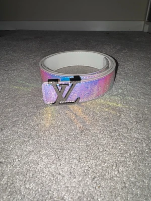 Holografiskt monogrambälte - Unikt bälte i holografisk finish med skimrande blå/lila ton. Dekorativt monogrammönster längs hela bältet och metallspänne med stilren bokstavsdesign. Ett riktigt statement-plagg som passar perfekt för att lyfta en enkel outfit. Passar både vardag och fest.