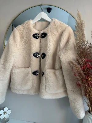 Beige teddyjacka med detaljer - Säljer en fluffig beige teddyjacka med stora fickor framtill och snygga svarta spännen som stängning. Jackan har rund halsringning och är helfodrad. 
