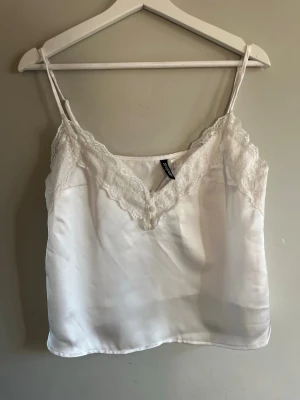 Vit spetsdetaljerad linnetopp H&M - Säljer en vit linnetopp från H&M Divided i storlek S. Toppen har tunna axelband, v-ringning med spetsdetaljer och små dekorativa knappar framtill. Materialet är glansigt och känns mjukt mot huden, perfekt för sommarens alla tillfällen.