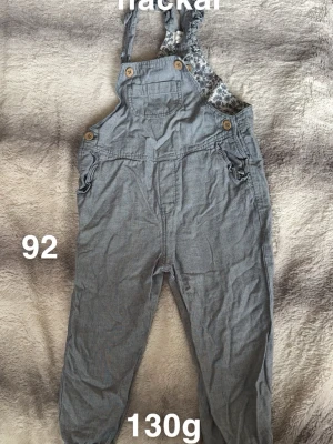 Blå byxdress med knappar, stl 92 - Säljer en blå byxdress med hängslen och träknappar. Byxdressen har en bröstficka och fina detaljer vid sidorna. Insidan upptill har ett blommigt mönster. Perfekt för dig som gillar en avslappnad och söt stil.🌸Fläckar. Storlek 92