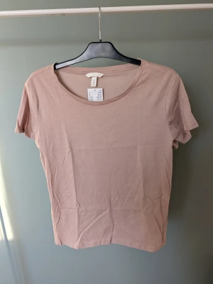 Beige basic t-shirt från H&M - Enkel och stilren beige t-shirt från H&M i mjuk bomull. Modellen har rund halsringning och korta ärmar, perfekt till alla typer av outfits. T-shirten har en clean look utan mönster eller detaljer.