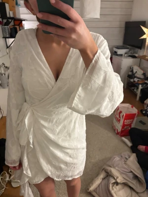 Vit omlottklänning från Gina Tricot - Supersnygg vit kort omlottklänning från Gina Tricot med långa ärmar och knyt i midjan. Klänningen har subtilt blommönster i tyget och v-ringad hals. Perfekt för dig som vill ha en fräsch och stilren look med lite extra detalj.