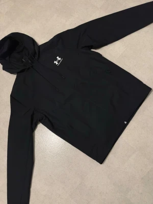 Svart vindjacka från Under Armour - Svart vindjacka från Under Armour med huva och dragsko. Jackan har en stilren design med logga på bröstet, hel dragkedja och meshfoder på insidan. Perfekt för aktiva dagar och sportiga outfits.