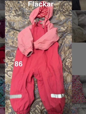 Rosa skaloverall med huva, stl 86 - Säljer en rosa skaloverall med ljusrosa ärmar och huva med rosa- och vitrandigt foder. Overallen har två dragkedjor framtill och reflexdetaljer på benen. Perfekt för kyliga dagar och lek utomhus.🌸 Fläckar. Storlek 86