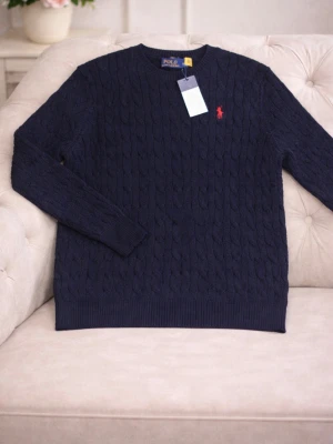 Marinblå stickad tröja Polo Ralph Lauren - Kabelstickad marinblå tröja från Polo Ralph Lauren med rund halsringning och röd broderad logga på bröstet. Tröjan har långa ärmar och ribbade muddar vid ärmslut och nederkant. Perfekt för lager-på-lager under kyliga dagar.