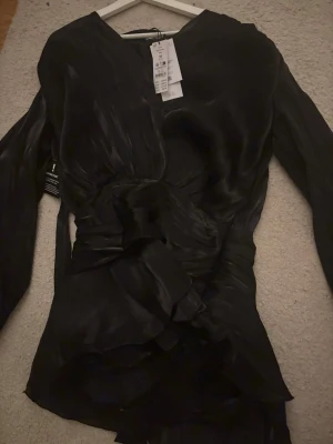 Svart miniklänning med volanger från Ginatricot  - Supersnygg svart miniklänning från ginatricot med v-ringning, långa ärmar och volangdetaljer. Klänningen är endast testad med prislapp kvar!