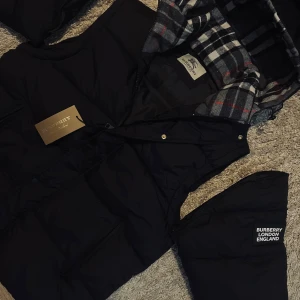Svart pufferjacka från Burberry - Snygg svart pufferjacka från Burberry med avtagbara ärmar så den kan bäras som väst. Jackan har huva med klassiskt rutigt foder i svart, vitt och rött samt dragkedja och tryckknappar framtill. Märkeslogga på ärmen och två praktiska fickor med dragkedja.