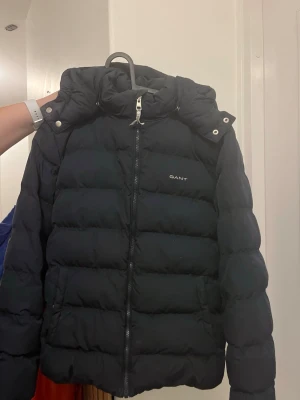 Svart pufferjacka från GANT - Säljer en svart pufferjacka från GANT med hög krage och avtagbar huva. Jackan har dragkedja framtill, tryckknappar vid kragen och GANT-logga broderad på bröstet samt längs dragkedjan. Perfekt för kalla dagar och riktigt snygg till alla outfits.
