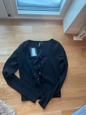Svart kofta med snörning från Pieces - Säljer en svart kofta från Pieces med långa ärmar och snygg snörning framtill. Koftan har en rak passform och är tillverkad i mjukt material som passar perfekt till lager på lager. Enkel men trendig design som funkar till många looks. Aldrig använts.