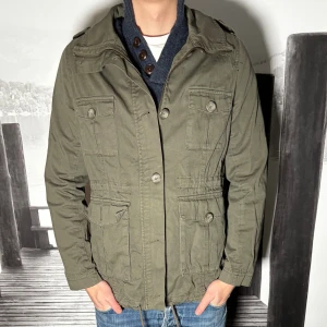 Olivgrön parkajacka från H&M - Säljer en olivgrön parkajacka från H&M med fyra stora fickor framtill, axelklaffar och knappar. Jackan har dragsko i midjan och huva, perfekt för dig som vill ha en stilren och funktionell jacka till vardags. Tillverkad i bomullsmix. 2 knappar av