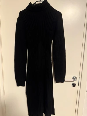 Svart ribbad stickad långklänning - Säljer en svart ribbad stickad långklänning med polokrage och långa ärmar. Klänningen har en slim passform och är perfekt för kyligare dagar. Enkel men ändå snygg design som är lätt att styla.
